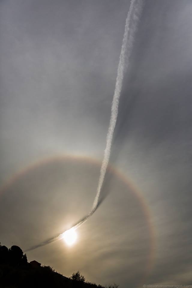 What's happened to the sky? Several common features of the daytime sky are interacting in uncommon ways.  First, well behind the silhouetted hills, is the typically bright Sun.  In front of the Sun are thin clouds, possibly the home to a layer of hexagonal ice crystals that together are creating the 22 degree halo of light surrounding the Sun. The unusual bent line that crosses the image is a contrail -- a type of cloud created by a passing airplane. Much of the contrail must actually be further away than the thin cloud because it casts a shadow onto the cloud, giving an unusual three-dimensional quality to the featured image. The featured image was taken in late January in the city of Patras in West Greece.