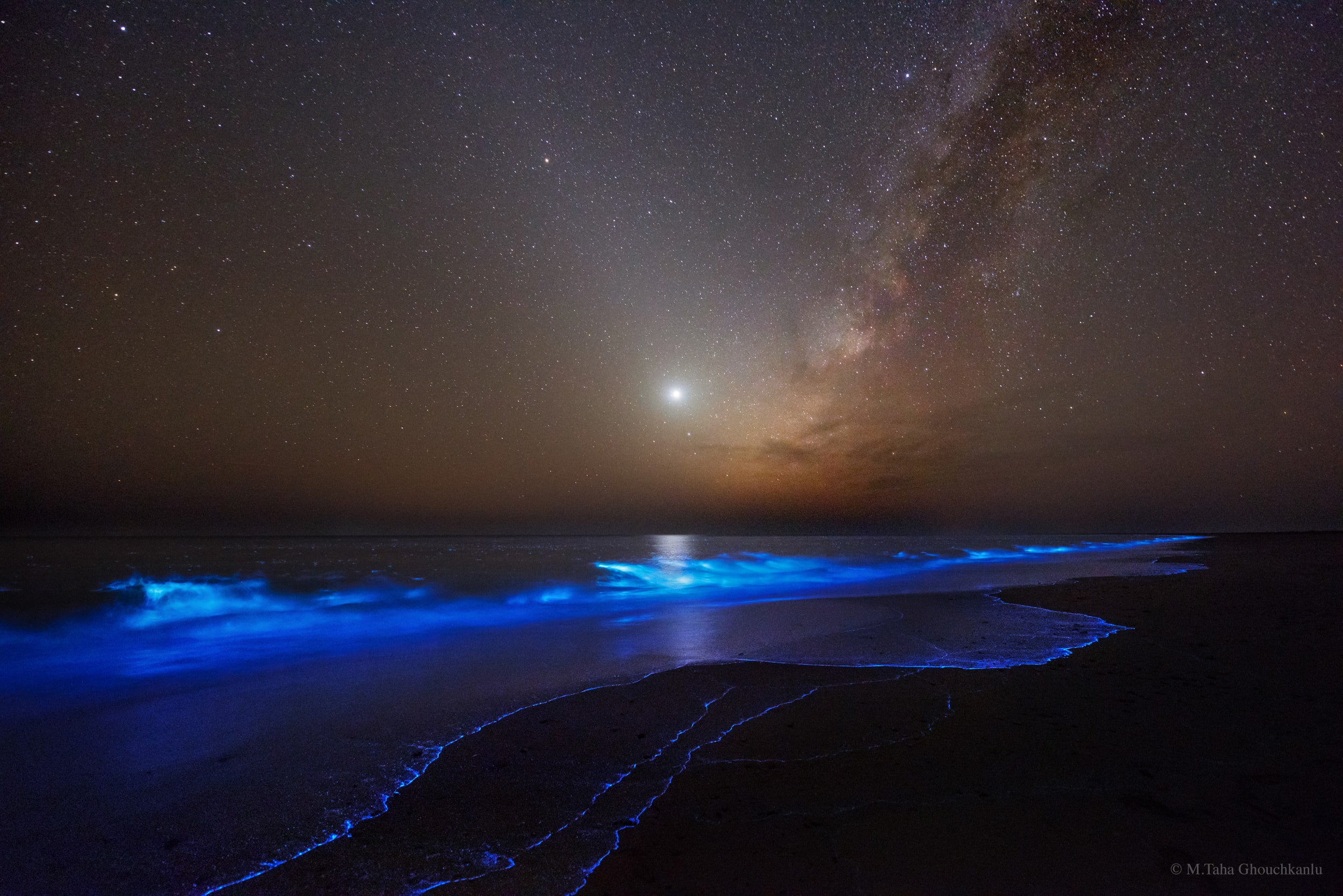 What glows in the night? This night, several unusual glows were evident -- some near, but some far. The foreground surf glimmers blue with the light of bioluminescent plankton.  Next out, Earth's atmosphere dims the horizon and provides a few opaque clouds. Farther out, the planet Venus glows bright near the image center. If you slightly avert your eyes, a diagonal beam of light will stand out crossing behind Venus. This band is zodiacal light, sunlight scattered by dust in our Solar System. Much farther away are numerous single bright stars, most closer than 100 light years away. Farthest away, also rising diagonally and making a "V" with the zodiacal light, is the central band of our Milky Way Galaxy. Most of the billions of Milky Way stars and dark clouds are thousands of light years away. The featured image was taken last November on the Iranian coast of Gulf of Oman.