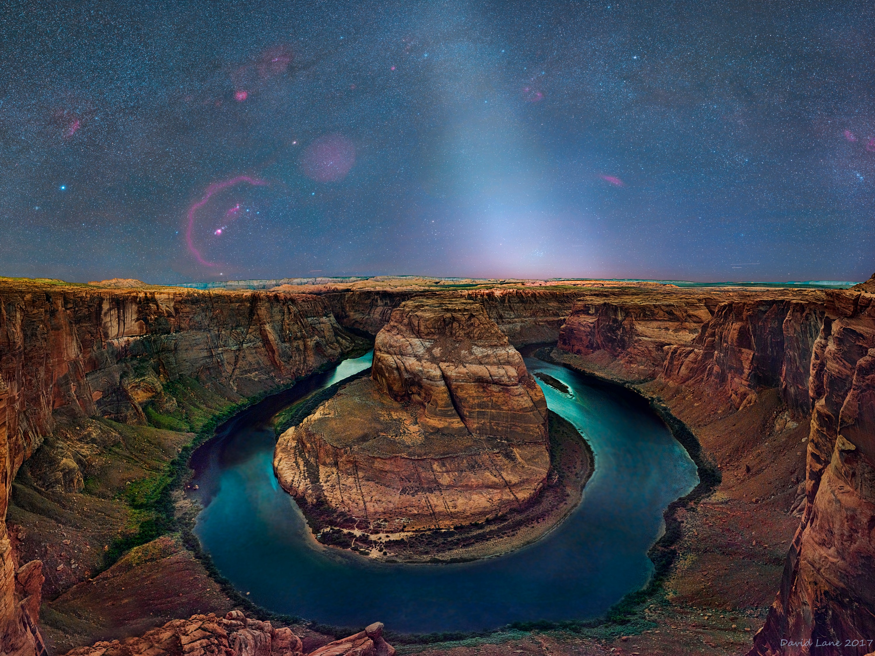 What's causing the unusual ray of white light extending upward from the central horizon? Dust orbiting the Sun. At certain times of the year, a band of sun-reflecting dust from the inner Solar System rises prominently before sunrise and is called zodiacal light.  The dust originates mostly from faint Jupiter-family comets and slowly spirals into the Sun. Pictured, in front of the zodiacal light, is a spectacular view of Horseshoe Bend of the Colorado River.  Emitted from well behind the zodiacal light is a spectacular sky that includes many bright stars including Sirius, several blue star clusters including the Pleiades, and an assortment of red nebulas including Barnard's Loop in Orion. The 30-image composite was taken earlier this month in nearly complete darkness only six inches from the edge of a dangerous cliff.