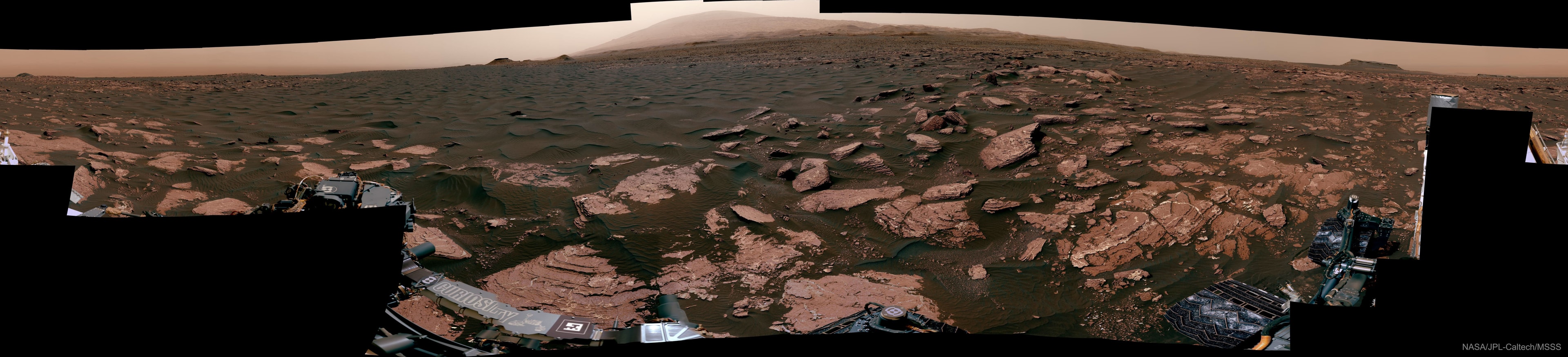 This was once a beach -- on ancient Mars. The featured 360-degree panorama, horizontally compressed, was taken by the robotic Curiosity rover currently exploring the red planet. Named Ogunquit Beach after its terrestrial counterpart, evidence shows that at times long ago the area was underwater, while at other times it was at the edge of an ancient lake.  The light peak in the central background is the top of Mount Sharp, the central feature in Gale Crater where Curiosity has been deployed. Curiosity is slowly ascending Mount Sharp.  Portions of the dark sands in the foreground have been scooped up for analysis.  The light colored bedrock is composed of sediment that likely settled at the bottom of the now-dried lakebed.  The featured panorama (interactive version here) was created from over 100 images acquired in late March and seemingly signed by the rover on the lower left.  Currently, Curiosity is carefully crossing deep megaripples of dark sands on its way to explore  Vera Rubin Ridge.    Explore the Universe: Random APOD Generator
