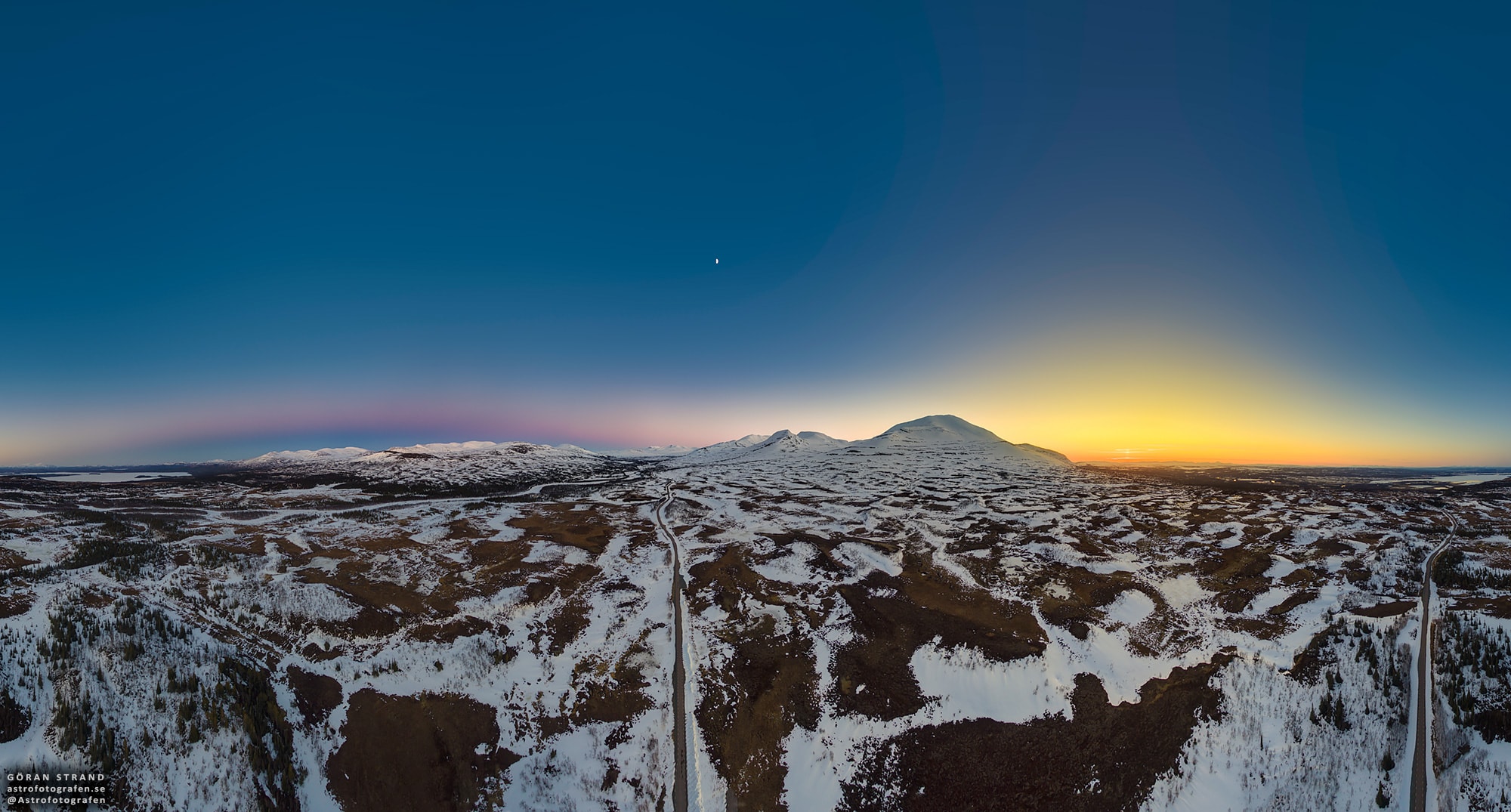 The road tracking through this stunning 360 degree view crosses a remote mountain area of Jämtland, Sweden, planet Earth. A 3x8 mosaic of still images, the panorama was taken on May 3rd from a small drone 200 meters above the ground. The scene records the warm light of the Sun setting in the northwest and the planet's dark shadow rising in the southeast. A small sun pillar gives away the Sun's position just below the horizon while the pinkish anti-twilight arch or belt of Venus outlines Earth's shadow. In the middle of the remarkably clear sky, the First Quarter Moon is visible above the mountains, its half illuminated disk separating shadowrise and sunset. A range of exposures was used to cover the large difference in sky brightness. The tallest peak left of the sunset is Storsnasen, some 1400 meters above sea level.