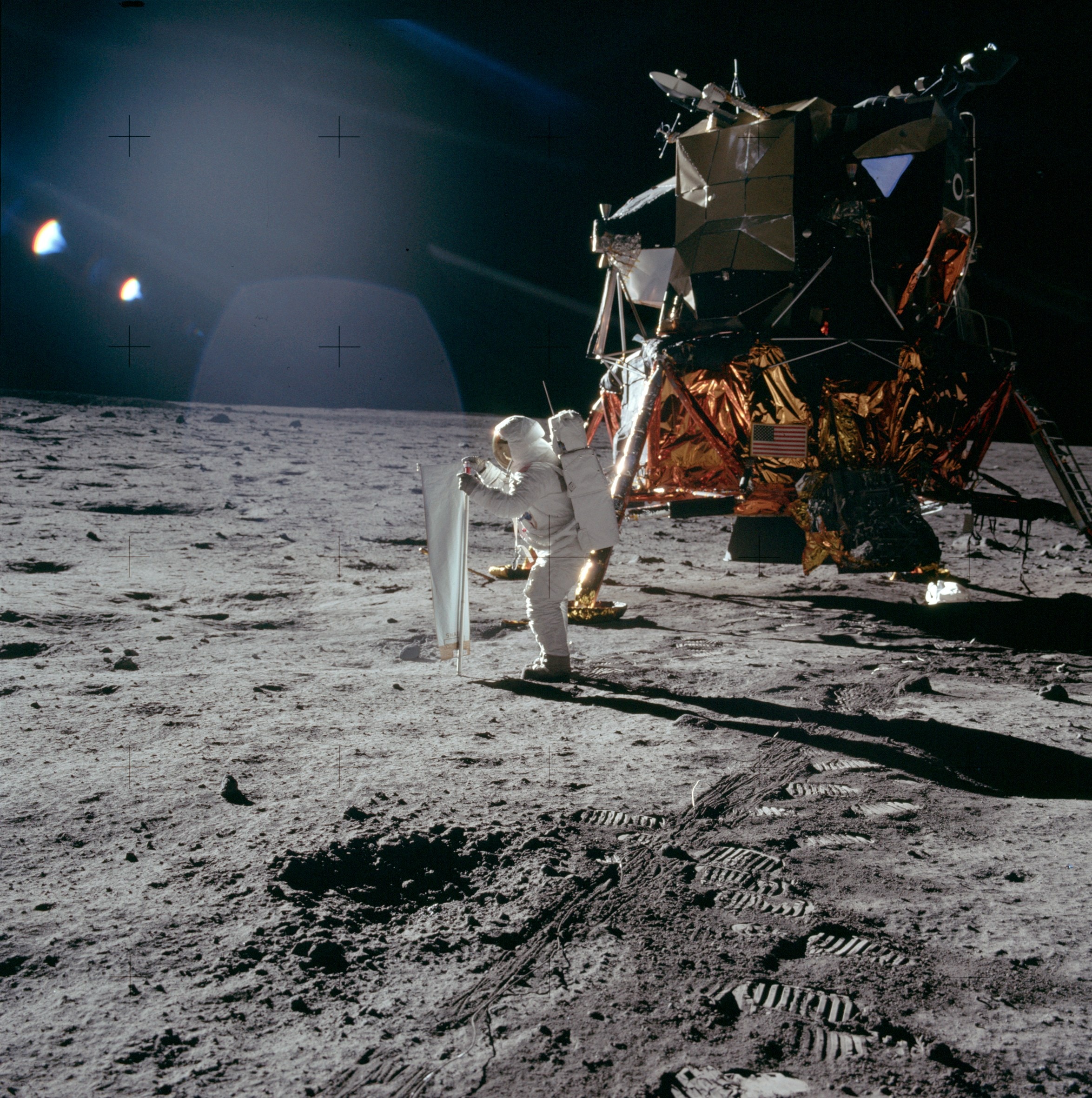 Bright sunlight glints and long dark shadows mark this image of the lunar surface. It was taken July 20, 1969 by Apollo 11 astronaut Neil Armstrong, the first to walk on the Moon. Pictured is the mission's lunar module, the Eagle, and spacesuited lunar module pilot Buzz Aldrin unfurling a long sheet of foil also known as the Solar Wind Composition Experiment. Exposed facing the Sun, the foil trapped particles streaming outward in the solar wind, catching a sample of material from the Sun itself. Along with moon rocks and lunar soil samples, the solar wind collector was returned for analysis in earthbound laboratories.