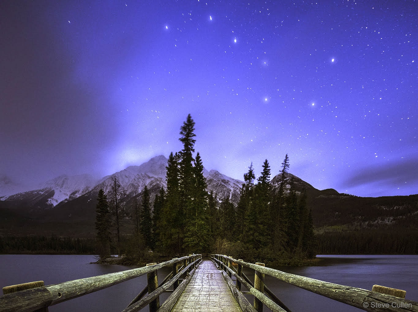 When did you first learn to identify this group of stars? Although they are familiar to many people around the world, different cultures have associated this asterism with different icons and folklore. Known in the USA as the Big Dipper, the stars are part of a constellation designated by the International Astronomical Union in 1922 as the Great Bear (Ursa Major).  The recognized star names of these stars are (left to right) Alkaid, Mizar/Alcor, Alioth, Megrez, Phecda, Merak, and Dubhe.  Of course, stars in any given constellation are unlikely to be physically related. But surprisingly, most of the Big Dipper stars do seem to be headed in the same direction as they plough through space, a property they share with other stars spread out over an even larger area across the sky.  Their measured common motion suggests that they all belong to a loose, nearby star cluster, thought to be on average only about 75 light-years away and up to 30 light-years across. The cluster is more properly known as the Ursa Major Moving Group. The featured image captured the iconic stars recently above Pyramid Mountain in Alberta, Canada.   Free APOD Lectures: January 5 in NYC & January 9 in DC