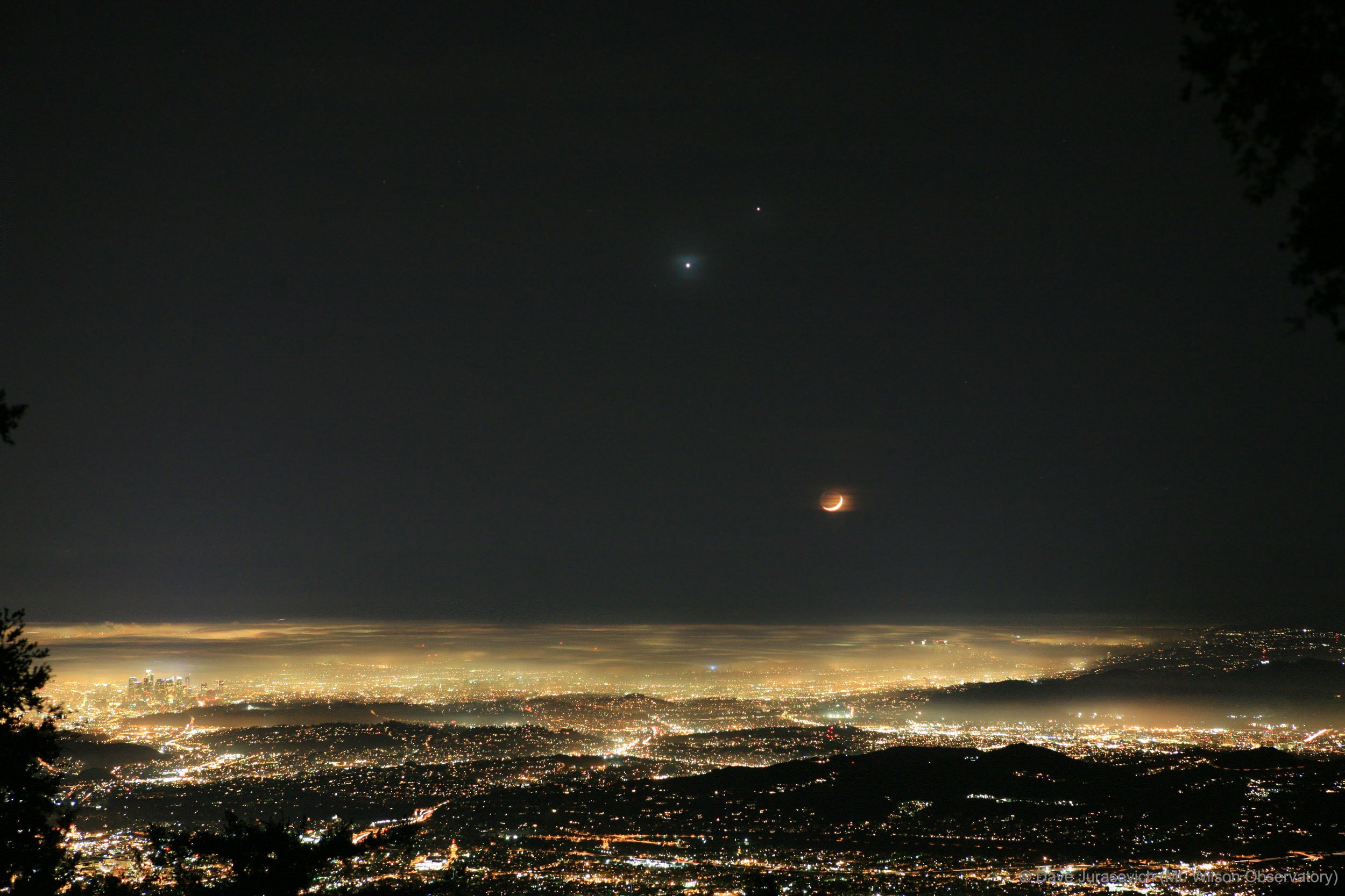 Sometimes, the sky may seem to smile over much of planet Earth.  On this day in 2008, visible the world over, was an unusual superposition of our Moon and the planets Venus and Jupiter.  Pictures taken at the right time show a crescent Moon that appears to be a smile when paired with the planetary conjunction of seemingly nearby Jupiter and Venus.  Pictured here is the scene as it appeared from Mt. Wilson Observatory overlooking Los Angeles, California, USA after sunset on 2008 November 30.  Highest in the sky and farthest in the distance is the planet Jupiter.  Significantly closer and visible to Jupiter's lower left is Venus, appearing through Earth's atmospheric clouds as unusually blue.  On the far right, above the horizon, is our Moon, in a waxing crescent phase.  Thin clouds illuminated by the Moon appear unusually orange.   Sprawling across the bottom of the image are the hills of Los Angeles, many covered by a thin haze, while LA skyscrapers are visible on the far left.  Hours after the taking of this image, the Moon approached the distant duo, briefly eclipsed Venus, and then moved on.  This week, another conjunction of Venus and Jupiter is occurring and is visible to much of planet Earth to the east just before sunrise.