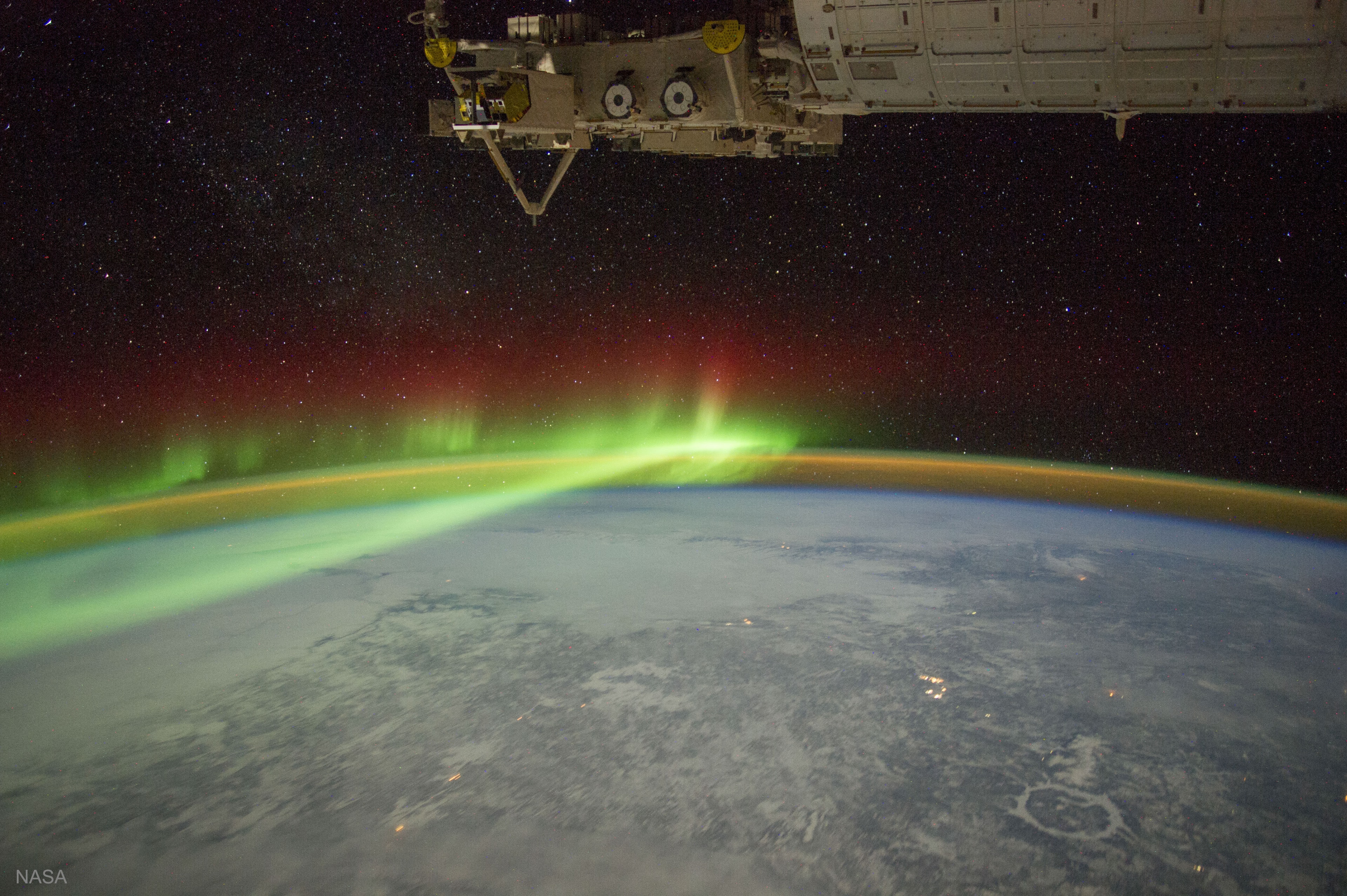 How many of these can you find in today's featured photograph: an aurora, airglow, one of the oldest impact craters on the Earth, snow and ice, stars, city lights, and part of the International Space Station? Most of these can be identified by their distinctive colors. The aurora here appears green at the bottom, red at the top, and is visible across the left of image. Airglow appears orange and can be seen hovering over the curve of the Earth. The circular Manicouagan Crater in Canada, about 100 kilometers across and 200 million years old, is visible toward the lower right and is covered in white snow and ice.  Stars, light in color, dot the dark background of space. City lights appear a bright yellow and dot the landscape. Finally, across the top, part of the International Space Station (ISS) appears mostly tan. The featured image was taken from the ISS in 2012.