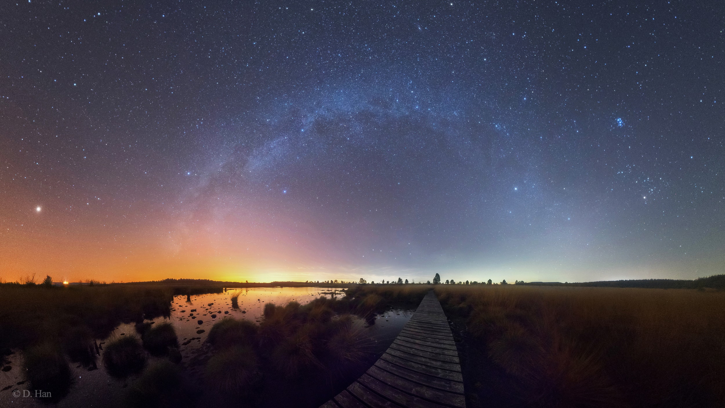 Taken near local midnight, this autumn night's panorama follows the arch of the Milky Way across the northern horizon from the High Fens, Eifel Nature Park at the border of Belgium and Germany. Shift your gaze across the wetlands from west to east (left to right) and you can watch stars once more prominent in northern summer give way to those that will soon dominate northern winter nights. Setting, wanderer Mars is brightest at the far left, still shining against almost overwhelming city lights along the southwestern horizon. Bright stars Altair, Deneb, and Vega form the northern sky's summer triangle, straddling the Milky Way left of center. Part of the winter hexagon Capella and Aldebaran, along with the beautiful Pleiades star cluster shine across the northeastern sky. The line-of-sight along the hikers boardwalk leads almost directly toward the Big Dipper, an all season asterism from these northern latitudes. Follow the Big Dipper's pointer stars to Polaris and the north celestial pole nearly centered above it. Andromeda, the other large galaxy in the skyscape, is near the top of the frame.