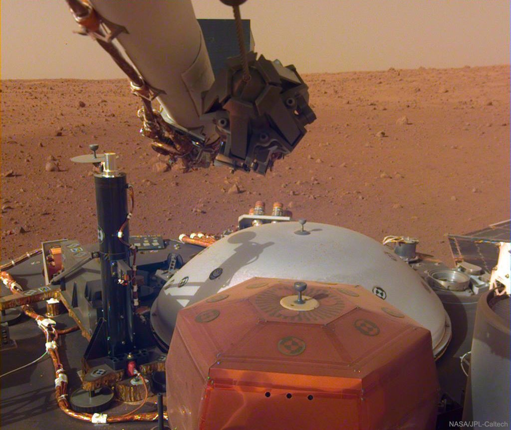 Your arm on Mars has unusual powers.  For one thing it is nearly 2 meters long, has a scoop and grapple built into its hand, and has a camera built into its forearm.  For another, it will soon deploy your ear -- a sensitive seismometer that will listen for distant rumblings -- onto the surface of Mars.  Your SEISmomet-ear is the orange box in the foreground, while the gray dome behind it will be its protective cover.  Your arm is attached to the InSight robotic lander that touched down on Mars two weeks ago. Somewhat unexpectedly, your ear has already heard something -- slight vibrations caused by the Martian wind flowing over the solar panels.  Light from the Sun is being collected by the solar panels, part of one being visible on the far right. Actually, at the present time, you have two arms operating on Mars, but they are separated by about 600 kilometers. That's because your other active arm is connected to the Curiosity rover exploring a distant crater.  Taken a week ago, rusty soil and rocks are visible in the featured image beyond Insight, as well as the orange sky of Mars.   Follow APOD on: Instagram, Facebook,  Reddit, or Twitter