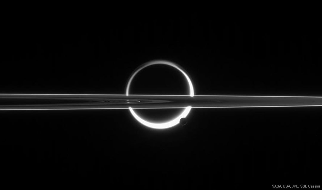 This is not a solar eclipse. Pictured here is a busy vista of moons and rings taken at Saturn. The large circular object in the center of the image is Titan, the largest moon of Saturn and one of the most intriguing objects in the entire Solar System. The dark spot in the center is the main solid part of the moon.  The bright surrounding ring is atmospheric haze above Titan, gas that is scattering sunlight to a camera operating onboard the robotic Cassini spacecraft. Cutting horizontally across the image are the rings of Saturn, seen nearly edge on.  At the lower right of Titan is Enceladus, a small moon of Saturn. Since the image was taken pointing nearly at the Sun, the surfaces of Titan and Enceladus appear in silhouette, and the rings of Saturn appear similar to a photographic negative. Now if you look really really closely at Enceladus, you can see a hint of icy jets shooting out toward the bottom of the image. It is these jets that inspired future proposals to land on Enceladus, burrow into the ice, and search for signs of extraterrestrial life.