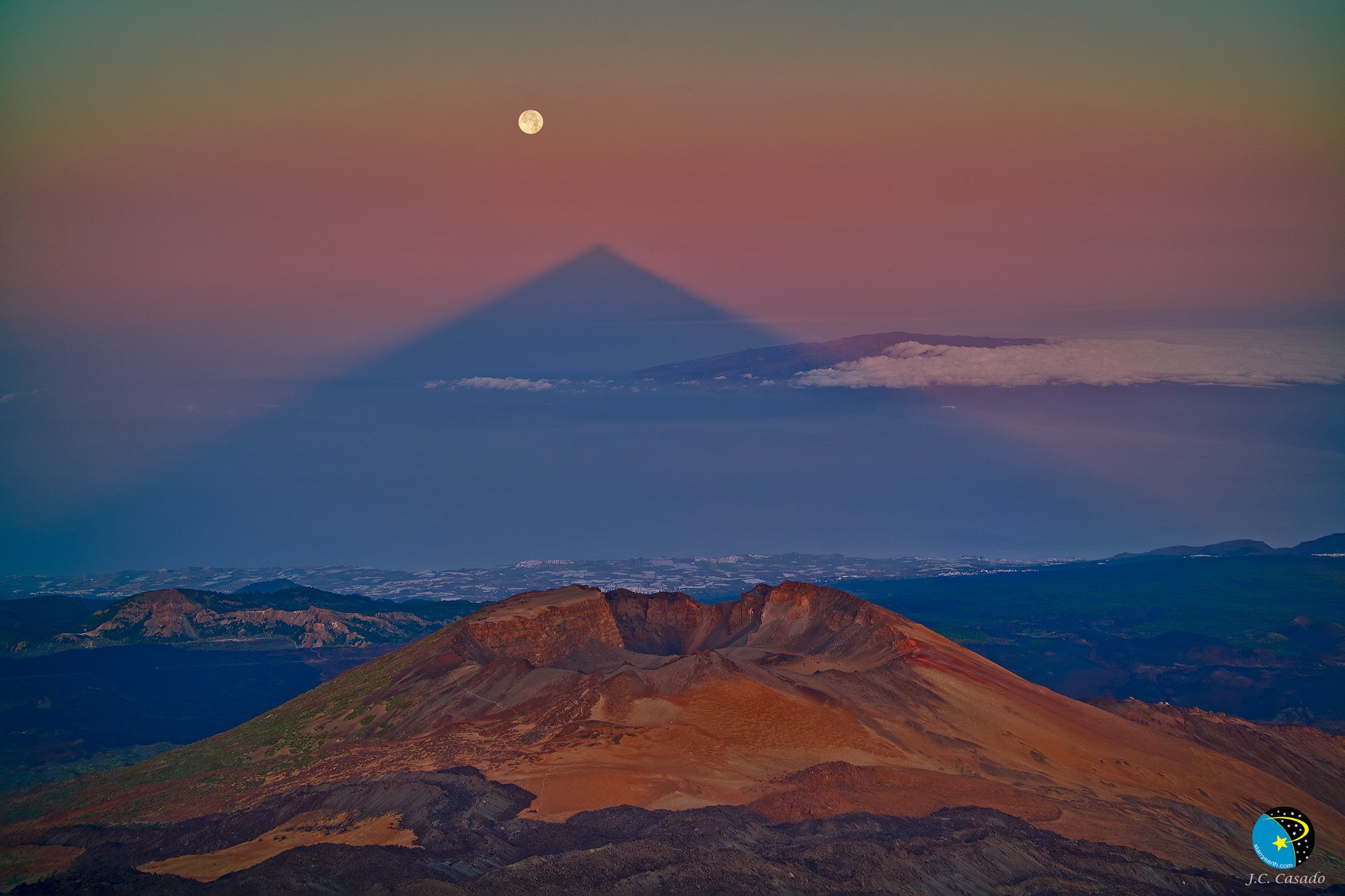 Why does the shadow of this volcano look like a triangle? The Mount Teide volcano itself does not have the strictly pyramidal shape that its geometric shadow might suggest.  The triangle shadow phenomena is not unique to the Mt. Teide, though, and is commonly seen from the tops of other large mountains and volcanoes. A key reason for the strange dark shape is that the observer is looking down the long corridor of a sunset (or sunrise) shadow that extends to the horizon. Even if the huge volcano were a perfect cube and the resulting shadow were a long rectangular box, that box would appear to taper off at its top as its shadow extended far into the distance, just as parallel train tracks do. The featured spectacular image shows Pico Viejo crater in the foreground, located on Tenerife in the Canary Islands of Spain. The nearly full moon is seen nearby shortly after its total lunar eclipse.   Explore the Universe: Random APOD Generator