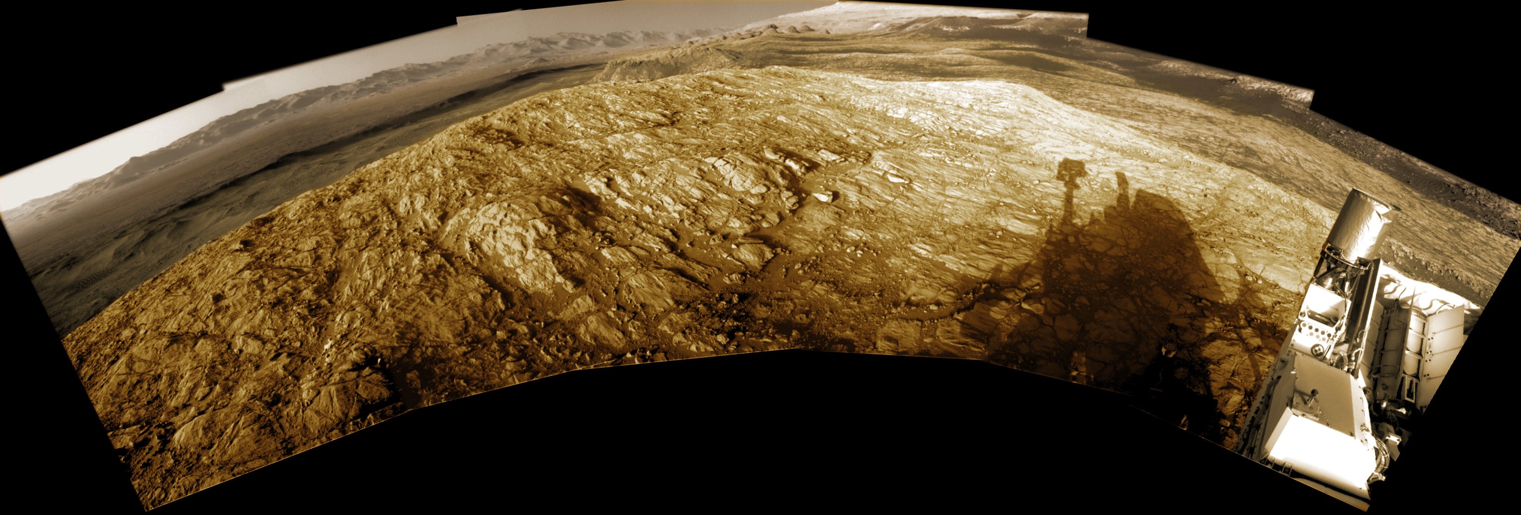 Shadows grow long near sunset in this wide panoramic view from the Curiosity rover on Mars. Made with Curiosity's navcam, the scene covers about 200 degrees from north through east to south (left to right), stitched together from frames taken by the Mars rover on sol 2616. That's just Earth date December 16. Curiosity is perched on top of a plateau on Western Butte. The distant northern rim of Gale crater is visible along the left. Near center is Central Butte, already visited by Curiosity. On the right, the shadow of the rover seems to stretch toward the base of Aeolis Mons (Mount Sharp), a future destination. The monochrome navcam frames have been colorized to approximate the colors of the late martian afternoon.