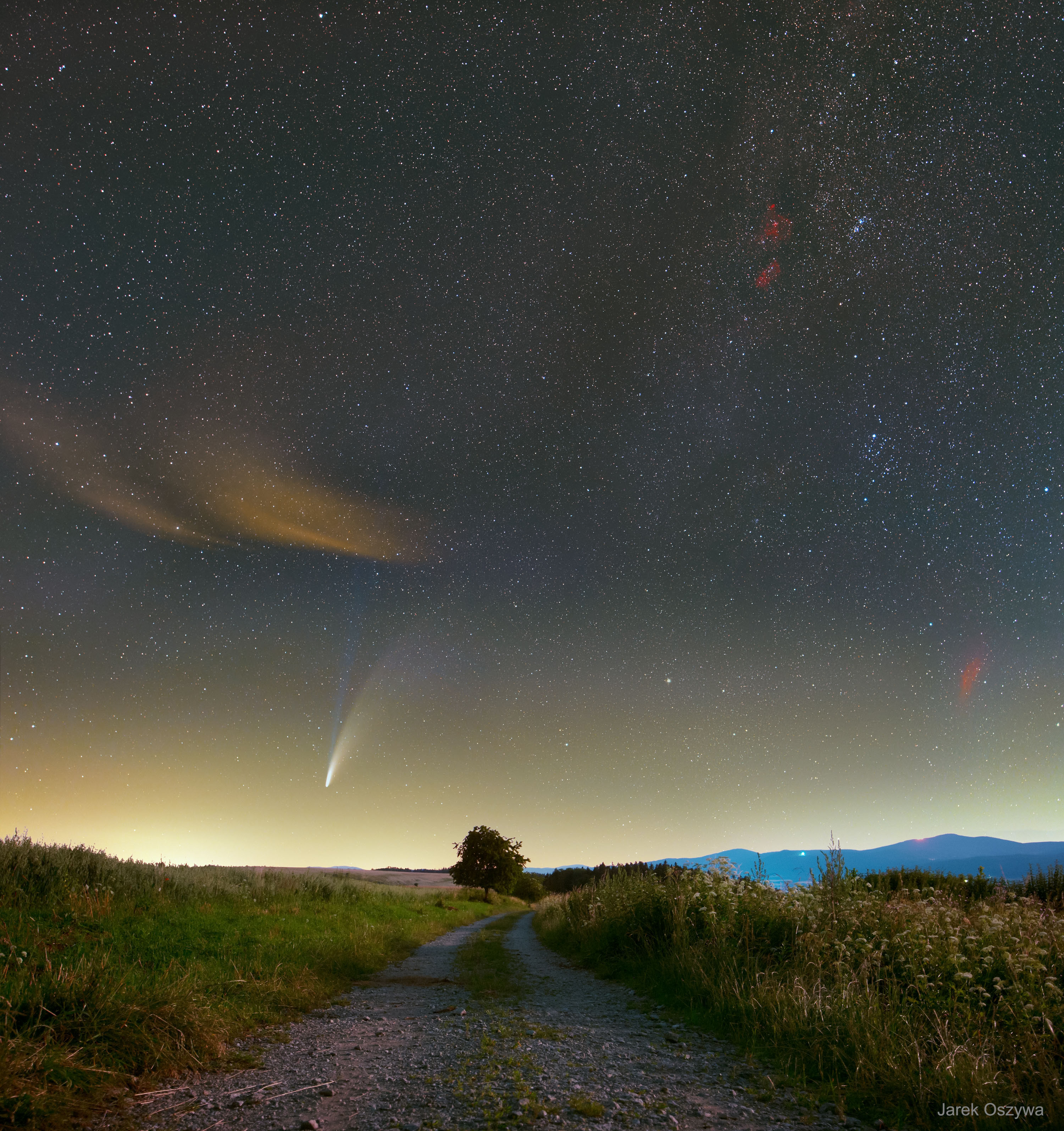 Would you brave wild animals to photograph this sky? One astrophotographer did -- and we all get to reap the rewards.  First, thousands of stars were visible with many of the brightest impressively blue. Next, several red-glowing nebulae were discernible, including the California Nebula on the far right, and, above it, the Heart and Soul nebulae. But the real reason to brave the local wildlife was Comet NEOWISE, visible on the left.  In the featured long-duration composite taken last week, Comet NEOWISE's blue-glowing ion tail points straight up, away from the rising Sun, while the Sun-reflecting dust tail trails off toward the right.  The picture combines three exposures taken consecutively over 10 minutes from the same location near Miedzygórze, Poland.  A moonlit dirt road shows the path ahead, while the Śnieznik Mountains is visible on the horizon. Comet C/2020 F3 (NEOWISE) passes its closest to the Earth next week, after which the 5-km wide, evaporating, icy dirtball will fade as it glides back to the outer Solar System.   Notable Images of Comet NEOWISE Submitted to APOD: July || 19  || 18   || 17   || 16   || 15  || 14  || 13  || 12  || 11  || 10 & earlier ||