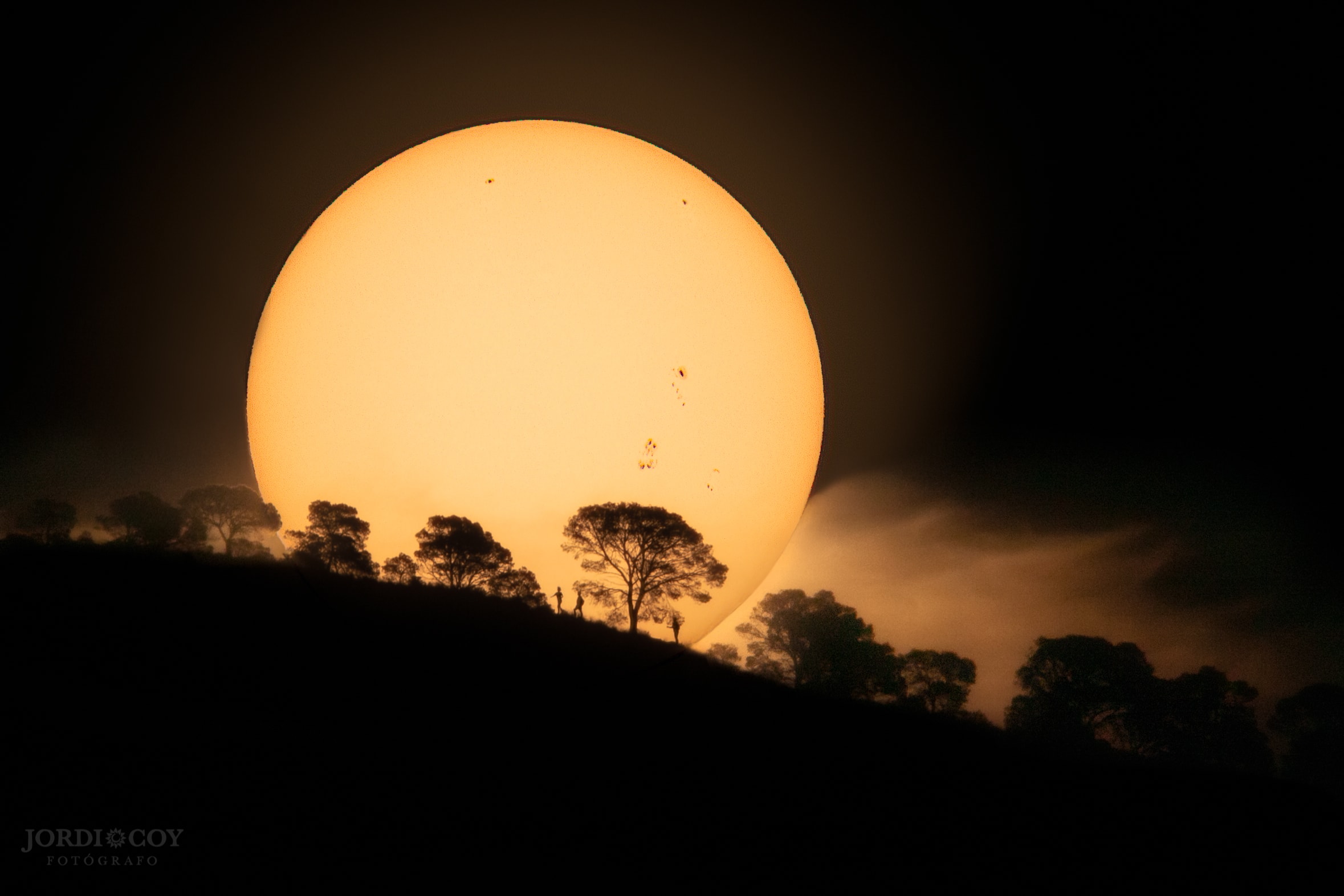 Is this giant orange ball about to roll down that tree-lined hill? No, because the giant orange ball is actually the Sun. Our Solar System's central star was captured rising beyond a hill on Earth twelve days ago complete with a delightfully detailed foreground.  The Sun's disk showed five sunspots, quite a lot considering that during the solar minimum in solar activity of the past few years, most days showed no spots. A close look at the hill -- Sierra del Cid in Perter, Spain -- reveals not only silhouetted pine trees, but silhouetted people -- by coincidence three brothers of the photographer.  The trees and brothers were about 3.5-kilometers away during the morning of the well-planned image. A dark filter muted the usually brilliant Sun and brought up great detail on the lower sunspots.  Within a few minutes, the Sun rose far above the hill, while within a week, the sunspots rotated around the Sun, out of view. The captured scene, however, is now frozen in time for all to enjoy.