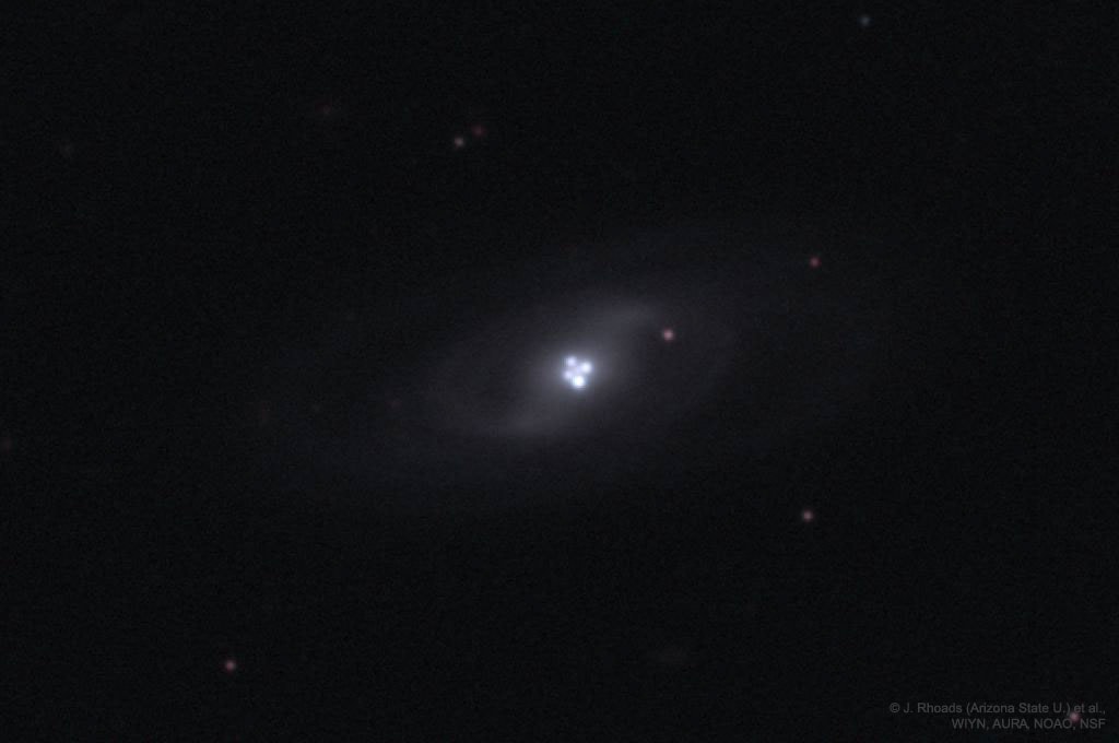 Most galaxies have a single nucleus -- does this galaxy have four?  The strange answer leads astronomers to conclude that the nucleus of the surrounding galaxy is not even visible in this image.  The central cloverleaf is rather light emitted from a background quasar.  The gravitational field of the visible foreground galaxy breaks light from this distant quasar into four distinct images.  The quasar must be properly aligned behind the center of a massive galaxy for a mirage like this to be evident.  The general effect is known as gravitational lensing, and this specific case is known as the Einstein Cross.  Stranger still, the images of the Einstein Cross vary in relative brightness, enhanced occasionally by the additional gravitational microlensing effect of specific stars in the foreground galaxy.