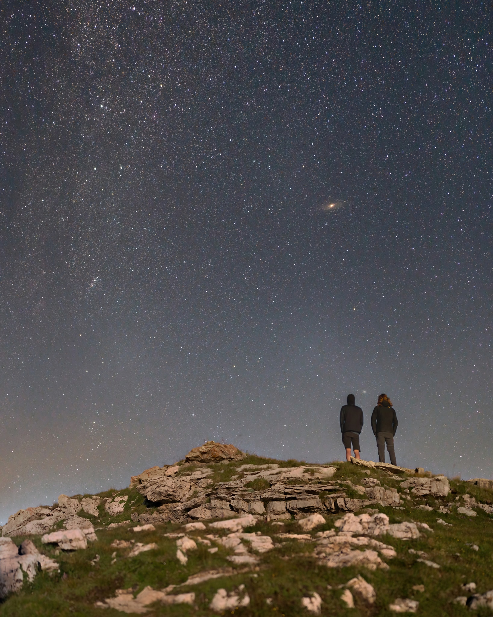 On an August night two friends enjoyed this view after a day's hike on the Plateau d'Emparis in the French Alps. At 2400 meters altitude the sky was clear. Light from a setting moon illuminates the foreground captured in the simple vertical panorama of images. Along the plane of our Milky Way galaxy stars of Cassiopeia and Perseus shine along the panorama's left edge. But seen as a faint cloud with a brighter core, the Andromeda galaxy, stands directly above the two friends in the night. The nearest large spiral galaxy, Andromeda is about 2.5 million light-years beyond the stars of the Milky Way. Adding to the evening's shared extragalactic perspective, the fainter fuzzy spot in the sky right between them is M33, also known as the Triangulum galaxy. Third largest in the local galaxy group, after Andromeda and Milky Way, the Triangulum galaxy is about 3 million light-years distant. On that night, the two friends stood about 3 light-nanoseconds apart.