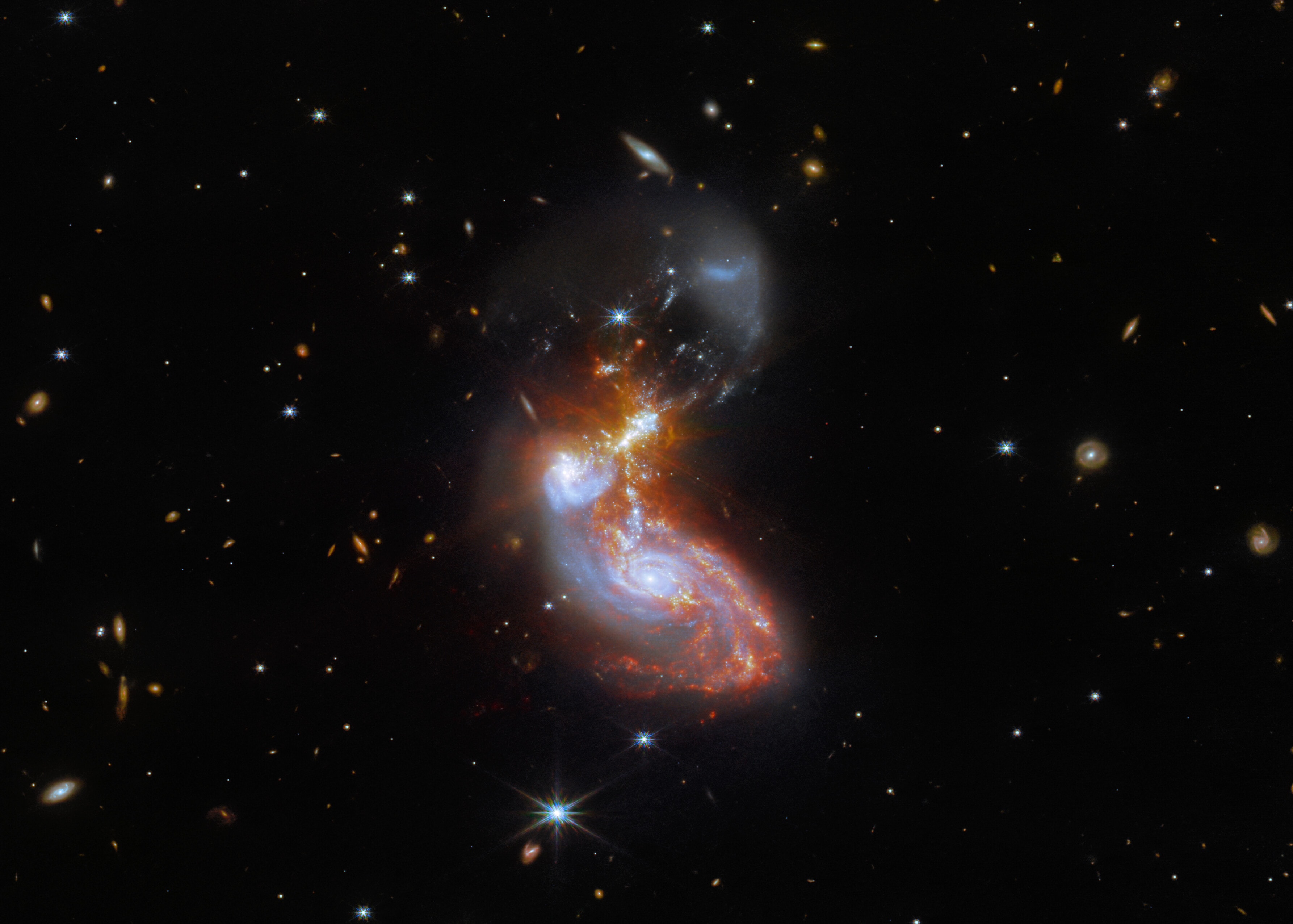 Bright at infrared wavelengths, this merging galaxy pair is some 500 million light-years away toward the constellation Delphinus. The cosmic mashup is seen against a background of even more distant galaxies, and occasional spiky foreground stars. But the galaxy merger itself spans about 100,000 light-years in this deep James Webb Space Telescope image. The image data is from Webb's Near-InfraRed Camera (NIRCam) and Mid-InfraRed Instrument (MIRI). Their combined, sharp infrared view follows galactic scale restructuring in the dusty merger's wild jumble of intense star forming regions  and distorted spiral arms