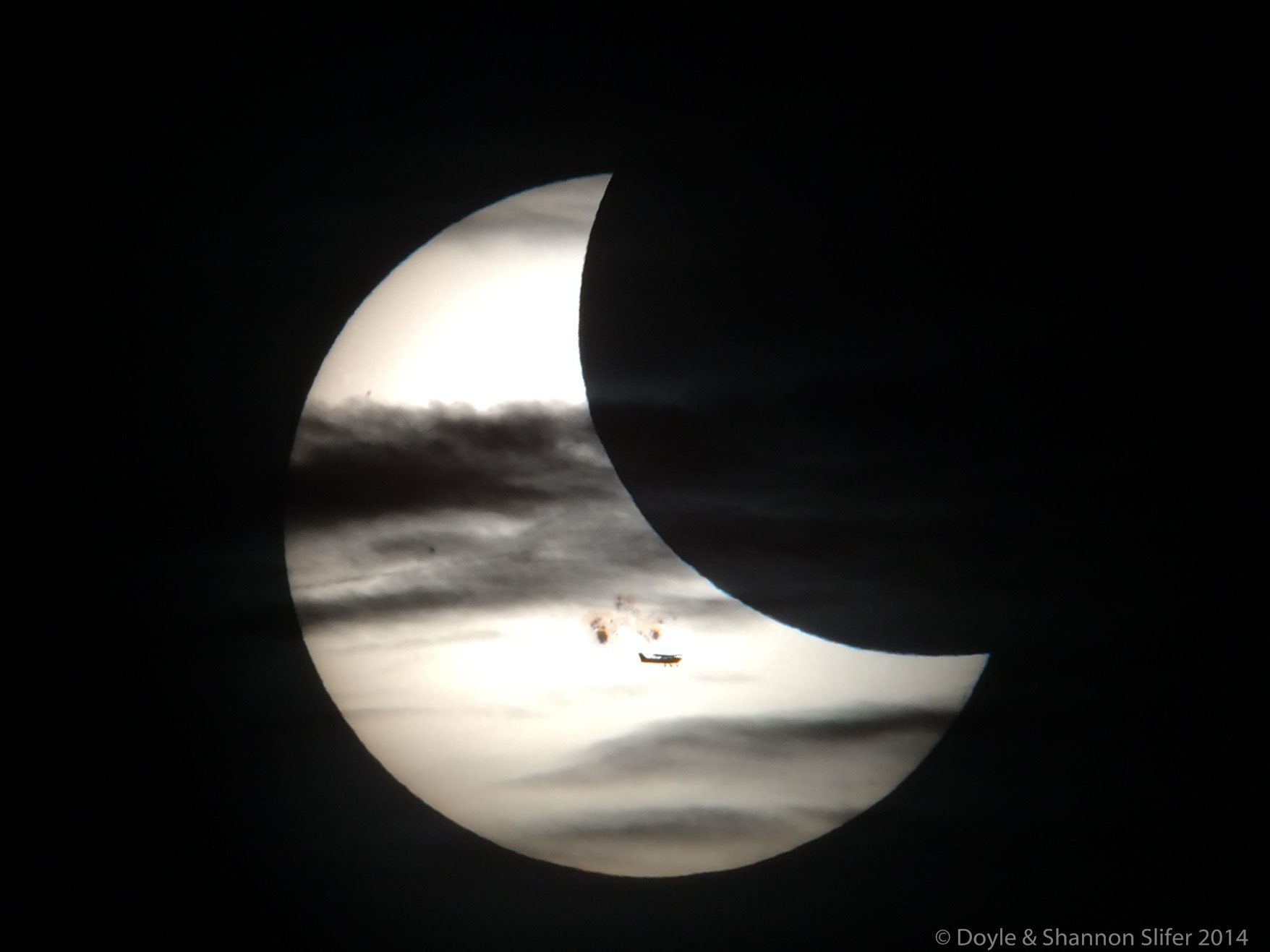 What's that in front of the Sun?  The closest object is an airplane, visible just below the Sun's center and caught purely by chance.  Next out are numerous clouds in Earth's atmosphere, creating a series of darkened horizontal streaks. Farther out is Earth's Moon, seen as the large dark circular bite on the upper right. Just above the airplane and just below the Sun's surface are sunspots. The main sunspot group captured here, AR 2192, was in 2014 one of the largest ever recorded and had been crackling and bursting with flares since it came around the edge of the Sun a week before. This show of solar silhouettes was unfortunately short-lived.  Within a few seconds the plane flew away. Within a few minutes the clouds drifted off. Within a few hours the partial solar eclipse of the Sun by the Moon was over. Fortunately, when it comes to the Sun, even unexpected  alignments are surprisingly frequent. Perhaps one will be imaged this Saturday when a new partial solar eclipse will be visible from much of North and South America.    APOD editor to speak: in Houghton, Michigan on Thursday, October 12 at 6 pm