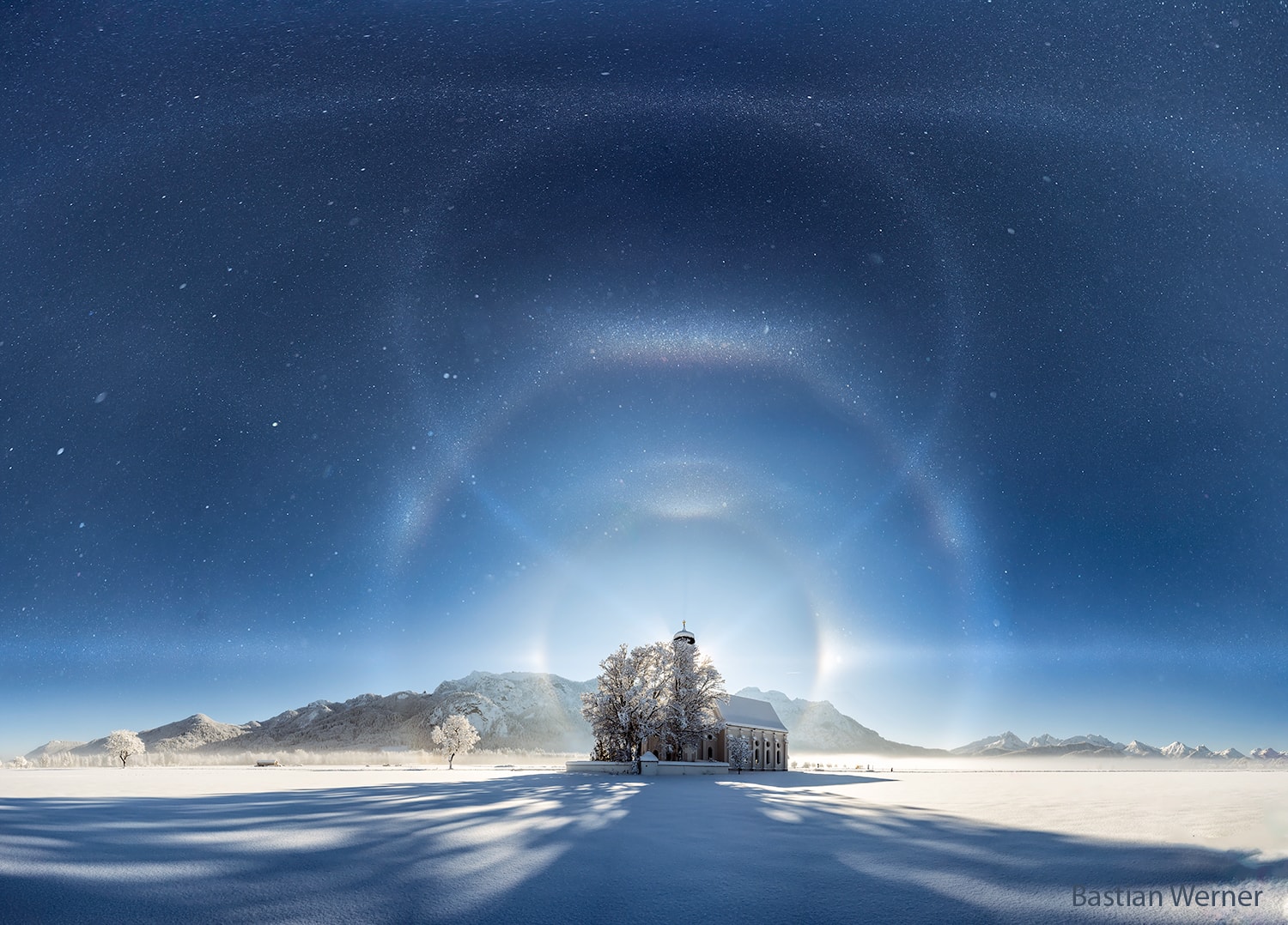 What's causing those unusual sky arcs? Ice crystals.  While crossing a field of fresh snow near Füssen, Bavaria, Germany, earlier this month, the photographer noticed that he had entered an ice fog.  For suspended water to freeze into an ice fog requires quite cold temperatures, and indeed the air temperature on this day was measured at well below zero.  The ice fog reflected light from the Sun setting behind St. Coleman Church.  The result was one of the greatest spectacles the photographer has ever seen. First, the spots in the featured picture are not background stars but suspended ice and snow. Next, two prominent ice halos are visible: the 22-degree halo and the 46-degree halo. Multiple arcs are also visible, including, from top to bottom, antisolar (subsun), circumzenithal, Parry, tangent, and parhelic (horizontal). Finally, the balloon shaped curve connecting the top arc to the Sun is the rarest of all: it is the heliac arc, created by reflection from the sides of hexagonally shaped ice crystals suspended in a horizontal orientation.