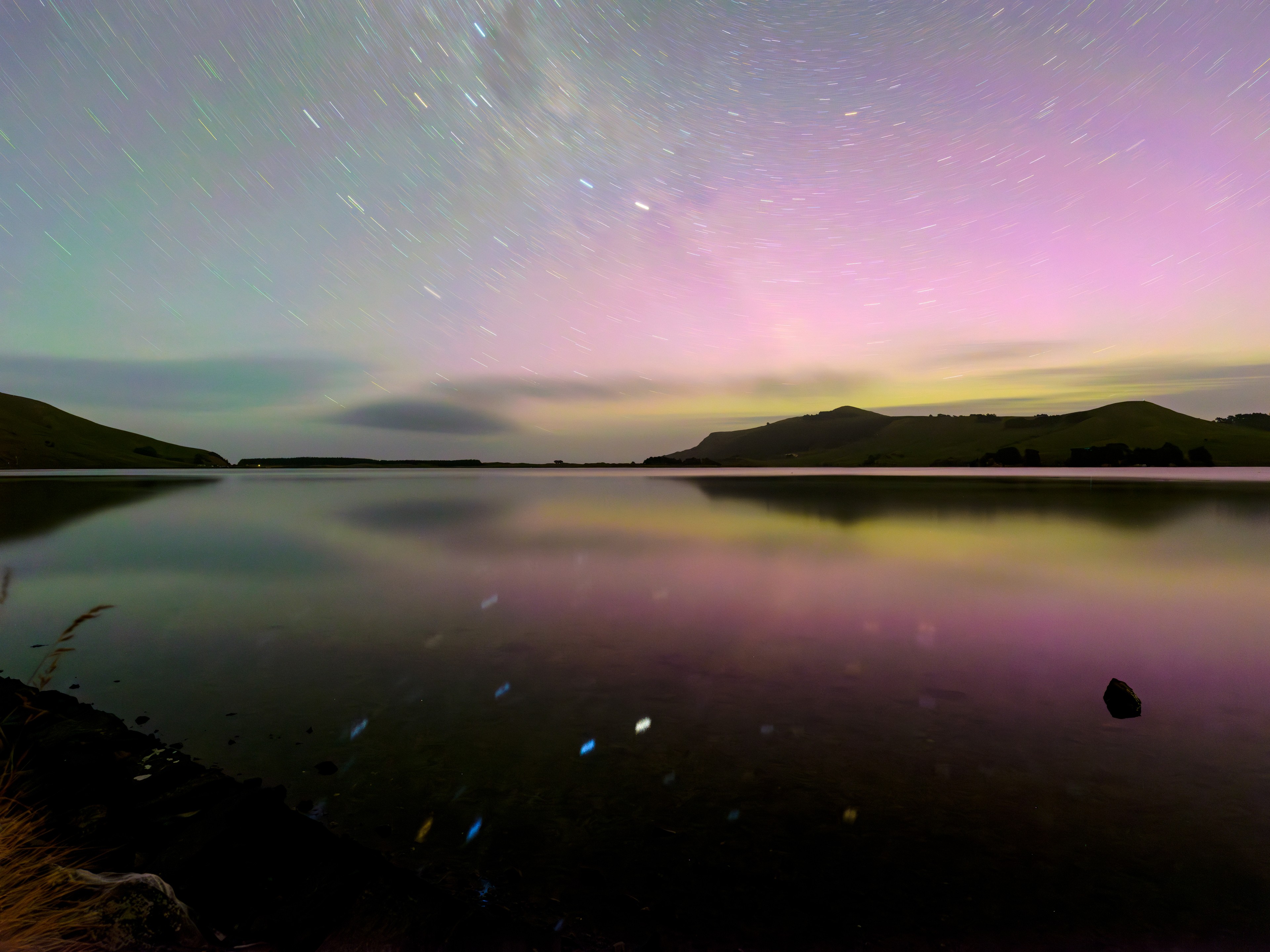 Colours of a serene evening sky are captured in this 8 minute exposure, made near this December's solstice from New Zealand, southern hemisphere, planet Earth. Looking south, star trails form the short concentric arcs around the rotating planet's south celestial pole positioned just off the top of the frame. At top and left of center are trails of the Southern Cross stars and a dark smudge from the Milky Way's Coalsack Nebula. Alpha and Beta Centauri make the brighter yellow and blue tinted trails, reflected below in the waters of Hoopers Inlet in the Pacific coast of the South Island's Otago Peninsula. On that short December summer night, aurora australis also gave luminous, green and reddish hues to the sky above the hills. Aurora shine as atoms and molecules in the upper atmosphere are excited by collisions with energetic particles. An upper atmospheric glow distinct from the aurora, a pale greenish airglow caused by a cascade of chemical reactions excited by sunlight, can be traced in diagonal bands at top left.