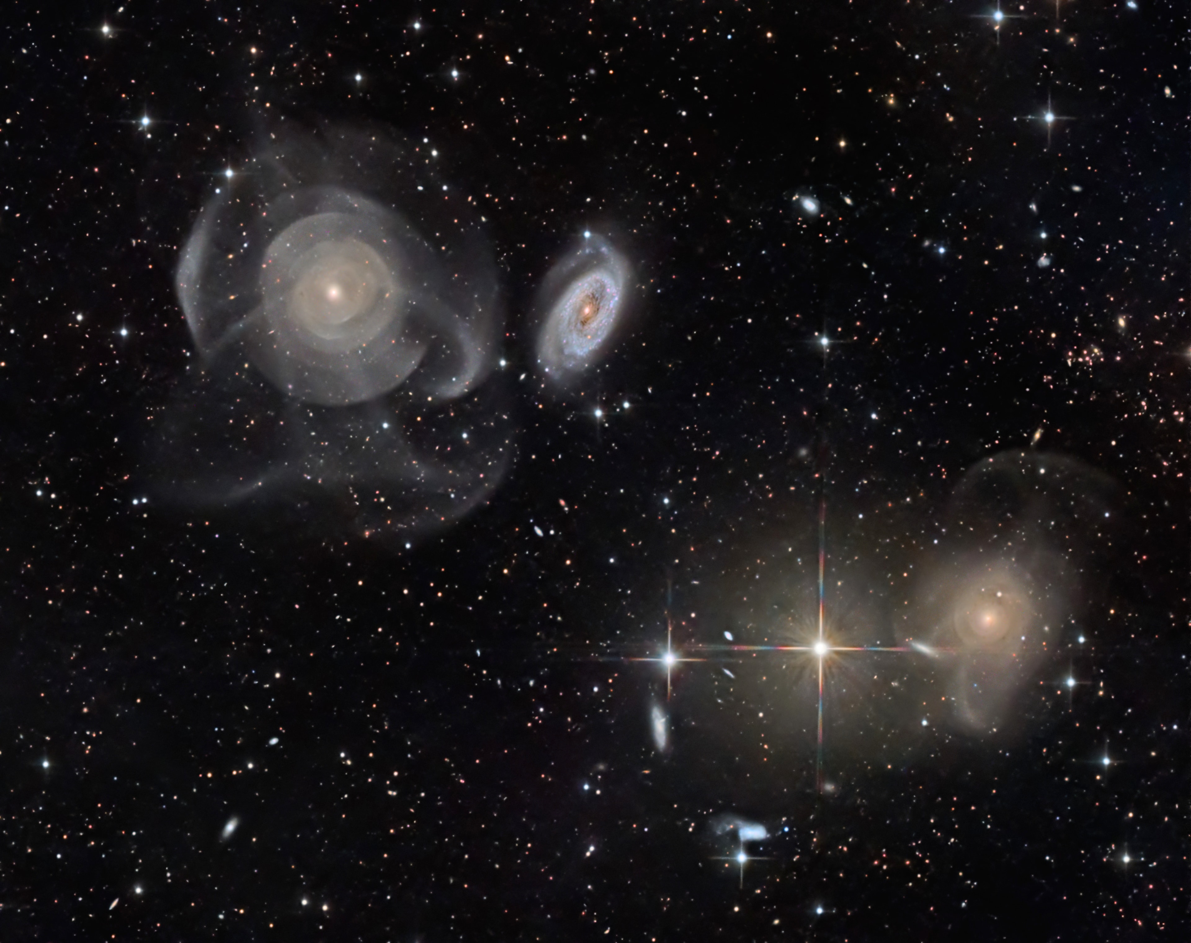 This spectacular intergalactic skyscape features Arp 227, a curious system of galaxies from the 1966 Atlas of Peculiar Galaxies. Some 100 million light-years distant within the boundaries of the constellation Pisces, Arp 227 consists of the two galaxies prominent above and left of center, the shell galaxy NGC 474 and its blue, spiral-armed neighbor NGC 470. The readily apparent shells and star streams of NGC 474 are likely tidal features originating from the accretion of another smaller galaxy during close gravitational encounters that began over a billion years ago. The large galaxy on the bottom righthand side of the deep image, NGC 467, appears to be surrounded by faint shells and streams too, evidence of another merging galaxy system. Intriguing background galaxies are scattered around the field that also includes spiky foreground stars. Of course, those stars lie well within our own Milky Way Galaxy. The telescopic field of view spans 25 arc minutes or just under 1/2 degree on the sky.