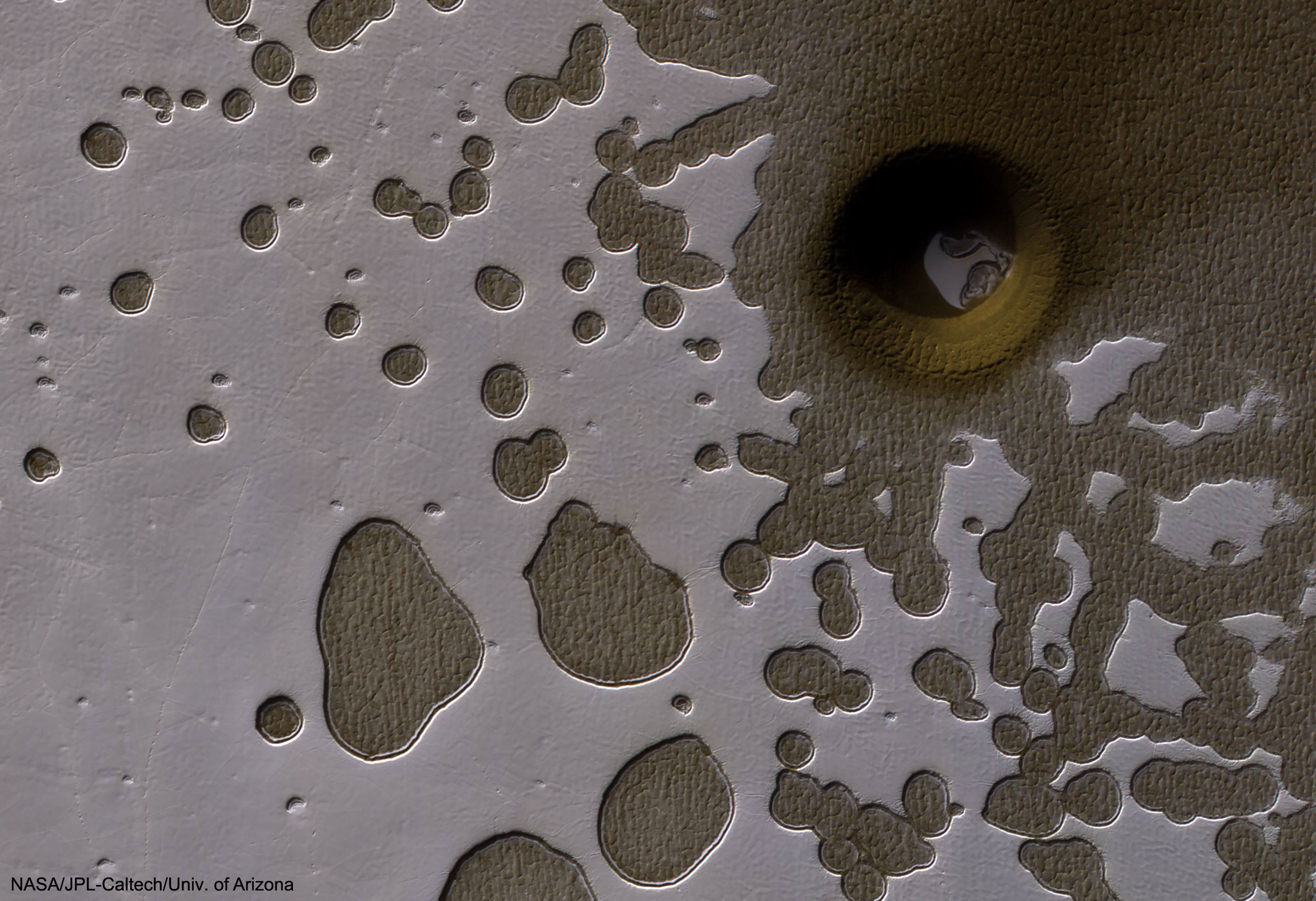 What created this unusual hole in Mars? Actually, there are numerous holes pictured in this Swiss cheese-like landscape, with all-but-one of them showing a dusty, dark, Martian terrain beneath evaporating, light, carbon-dioxide ice. The most unusual hole is on the upper right, spans about 100-meters, and seems to punch through to a lower level. Why this hole exists and why it is surrounded by a circular crater remains a topic of speculation, although a leading hypothesis is that it was created by a meteor impact. Holes such as this are of particular interest because they might be portals to lower levels that extend into expansive underground caves. If so, these naturally-occurring tunnels are relatively protected from the harsh surface of Mars, making them relatively good candidates to contain Martian life. These pits are therefore prime targets for possible future spacecraft, robots, and even human interplanetary explorers.