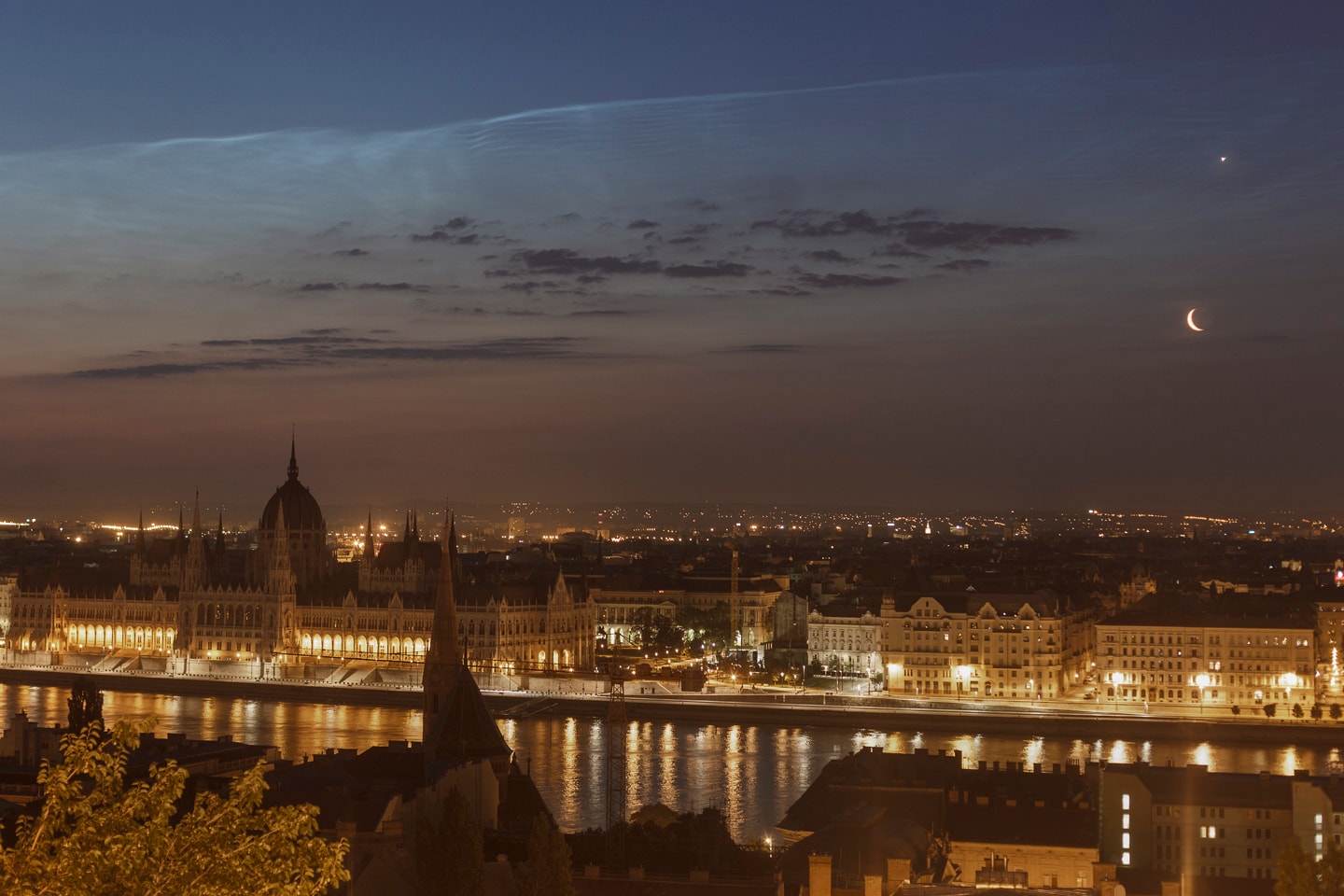 Before a solstice Sun rose on June 21, brilliant Venus and an old crescent Moon posed together over Budapest, Hungary for this predawn skyscape. In the foreground the view looks across the Danube river from Buda to Pest toward the dome and peaks of the Hungarian Parliament building. Low clouds are in silhouette against a twilight sky. But far enough above the eastern horizon to catch the sunlight shines another seasonal apparition on that solstice morning, noctilucent clouds. Seen near sunrise and sunset in summer months at high latitudes, the night-shining clouds are formed as water vapor in the cold upper atmosphere condenses on meteoric dust or volcanic ash near the edge of space.