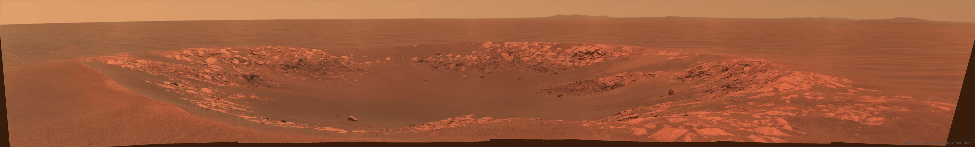 The robotic rover Opportunity sometimes passes small craters on Mars. Pictured here in 2010 is Intrepid Crater, a 20-meter across impact basin slightly larger than Nereus Crater that Opportunity had chanced across previously. The featured image is in approximately true color but horizontally compressed to accommodate a wide angle panorama. Intrepid Crater was named after the lunar module Intrepid that carried Apollo 12 astronauts to Earth's Moon 49 years ago. Beyond Intrepid Crater and past long patches of rusty Martian desert lie peaks from the rim of large Endeavour Crater, visible on the horizon. The Opportunity rover continues to explore Mars, recently surpassing 5,000 Martian days on the red planet.