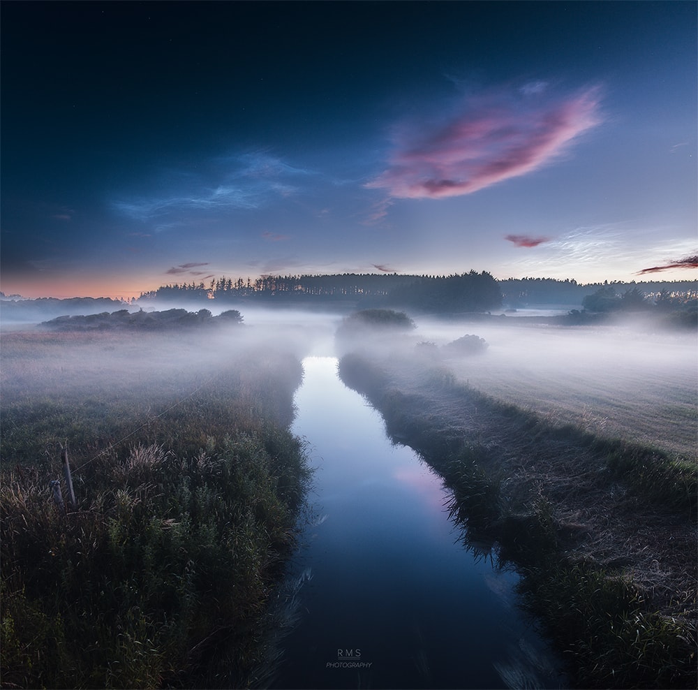 Near a summer's midnight a mist haunts the river bank in this dreamlike skyscape taken on July 3rd from northern Denmark. Reddened light from the Sun a little below the horizon gives an eerie tint to low hanging clouds. Formed near the edge of space, the silvery apparitions above them are noctilucent or night shining clouds. The icy condensations on meteoric dust or volcanic ash are still in full sunlight at the extreme altitudes of the mesophere. Usually seen at high latitudes in summer months, wide spread displays of the noctilucent clouds are now being reported.