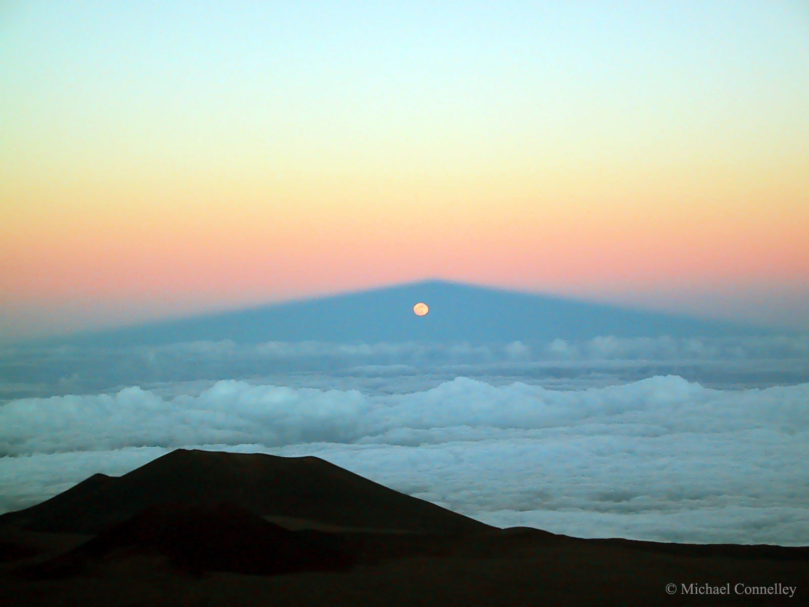 How can the Moon rise through a mountain? It cannot -- what was photographed here is a moonrise through the shadow of a large volcano. The volcano is Mauna Kea, Hawai'i, USA, a frequent spot for spectacular photographs since it is one of the premier observing locations on planet Earth. The Sun has just set in the opposite direction, behind the camera. Additionally, the Moon has just passed full phase -- were it precisely at full phase it would rise, possibly eclipsed, at the very peak of the shadow. The Moon is actually rising in the triangular shadow cone of the volcano, a corridor of darkness that tapers off in the distance like converging train tracks. The Moon is too large and too far away to be affected by the shadow of the volcano. Refraction of moonlight through the Earth's atmosphere makes the Moon appear slightly oval. Cinder cones from old volcanic eruptions are visible in the foreground.