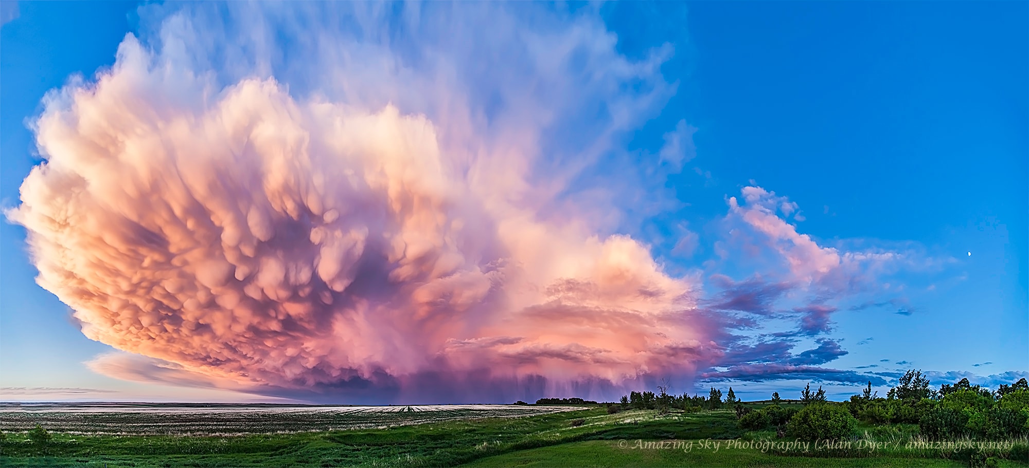 What type of cloud is that? This retreating cumulonimbus cloud, more commonly called a thundercloud, is somewhat unusual as it contains the unusual bumpiness of a mammatus cloud on the near end, while simultaneously producing falling rain on the far end. Taken in mid-2013 in southern Alberta, Canada, the cloud is moving to the east, into the distance, as the sun sets in the west, behind the camera. In the featured image, graphic sunset colors cross the sky to give the already photogenic cloud striking orange and pink hues. A darkening blue sky covers the background. Further in the distance, a rising, waxing, gibbous moon is visible on the far right.