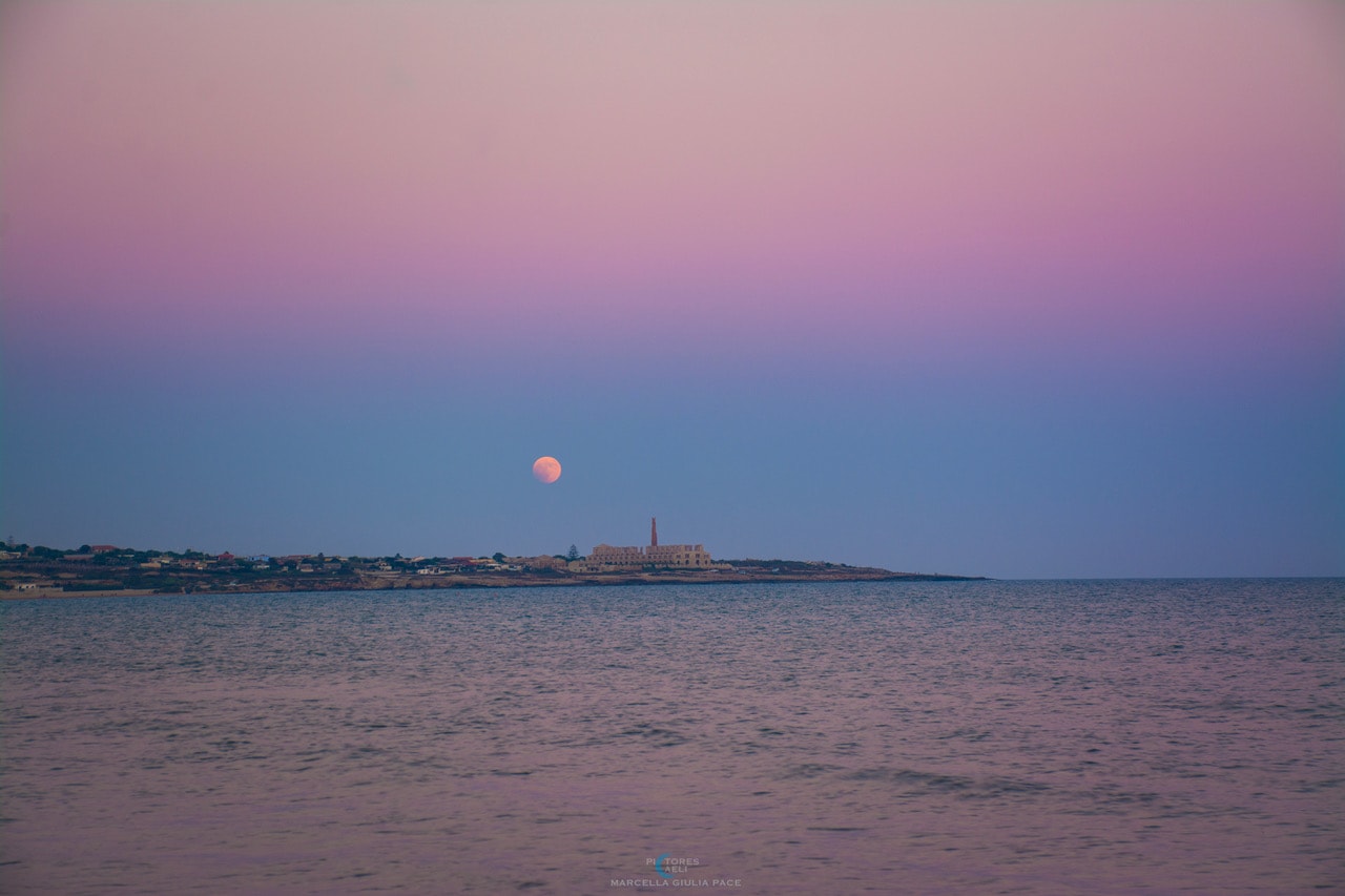 Can you find two Earth shadows in today's image? It's a bit tricky. To find the first shadow, observe that the top part of the atmosphere appears pink and the lower part appears blue. This is because the top half is exposed to direct sunlight, while the lower part is not. The purple area in between is known as the Belt of Venus, even though Venus can only appear on the other side of the sky, near the Sun. The blue color of the lower atmosphere is caused by the Earth blocking sunlight, creating Earth shadow number 1. Now, where is the second Earth shadow? Take a look at the Moon. Do you notice something unusual about the lower left part? That area appears unusually dark because it is in the shadow of the Earth, creating Earth shadow number 2. To be precise, the Moon was captured during a lunar eclipse. This carefully timed image was taken in Sampieri, Sicily, Italy, in July 2018.